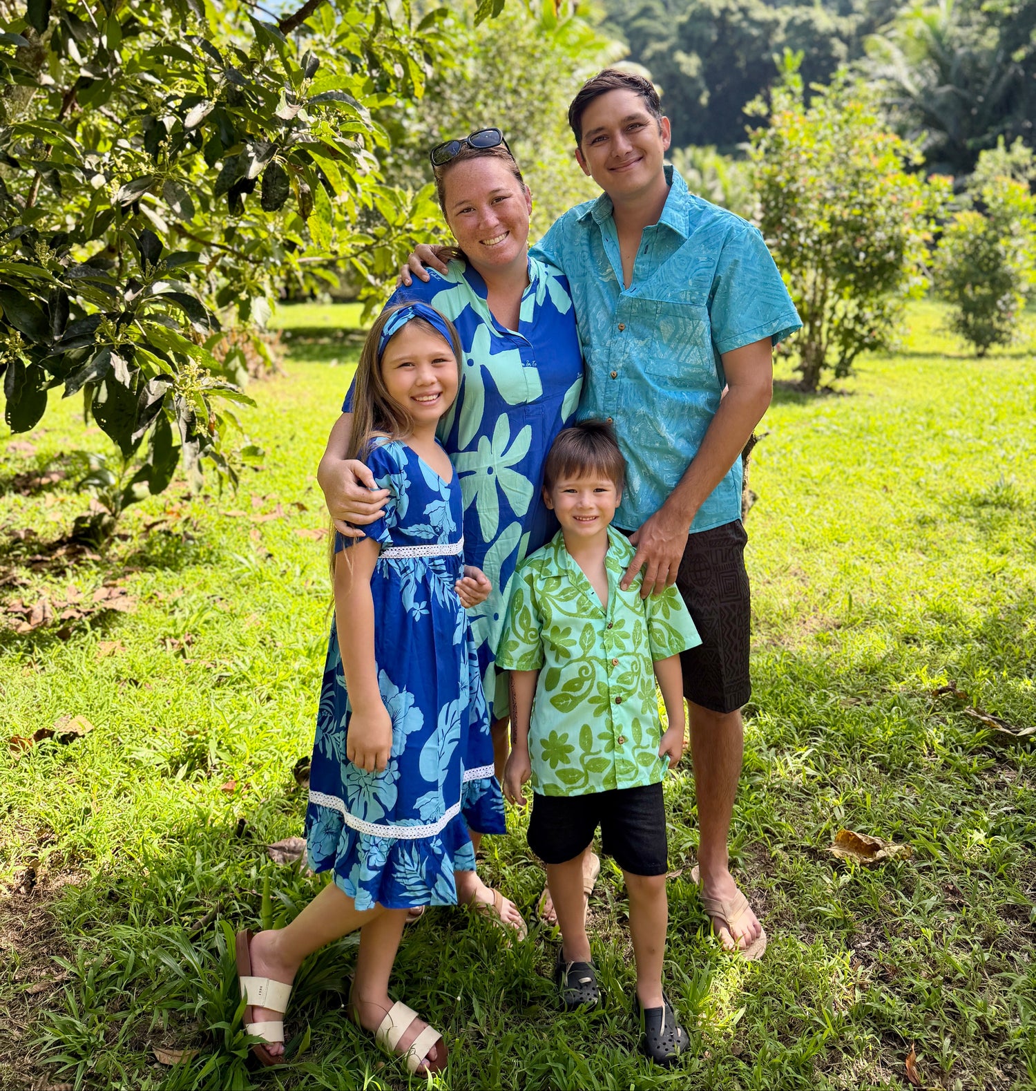 A family of four posing outdoors in a lush green orchard. The mother wears a blue floral dress and headband, the father wears a turquoise patterned short-sleeve button-up shirt and dark shorts, and the two children wear matching green and blue patterned shirts and shorts.