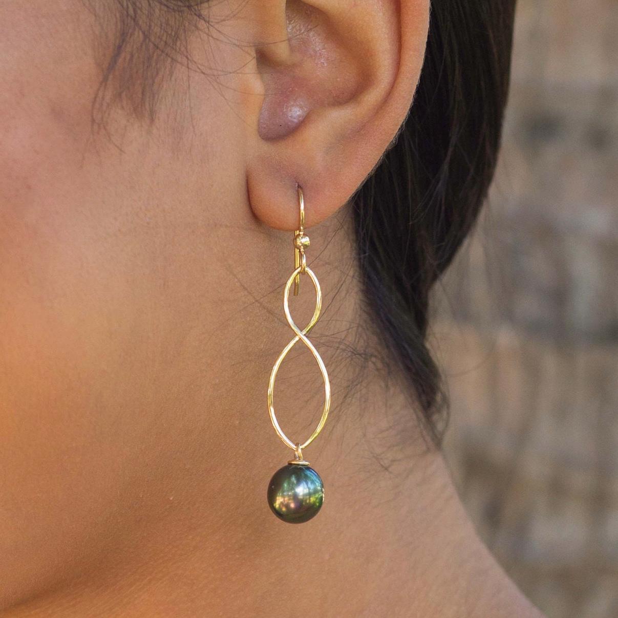 Gold-tone dangle earrings feature a twisted, infinity-like design.  A dark green, black Tahitian pearl is suspended at the bottom of each earring. The earrings have a simple, elegant style suitable for everyday wear or special occasions.